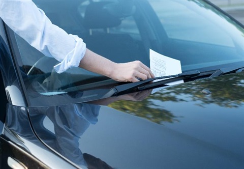 Person reaching for a parking fine placed under the wiper blade on a car