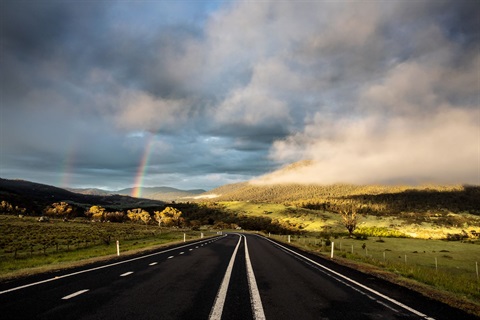 Alpine Way road surface and surrounding pastoral scenery, with a rainbow breaking through an overcast sky.