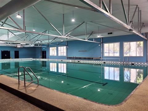 A photo of the renovated pool, inside, looking across the water's surface to the grounds outside through the windows.