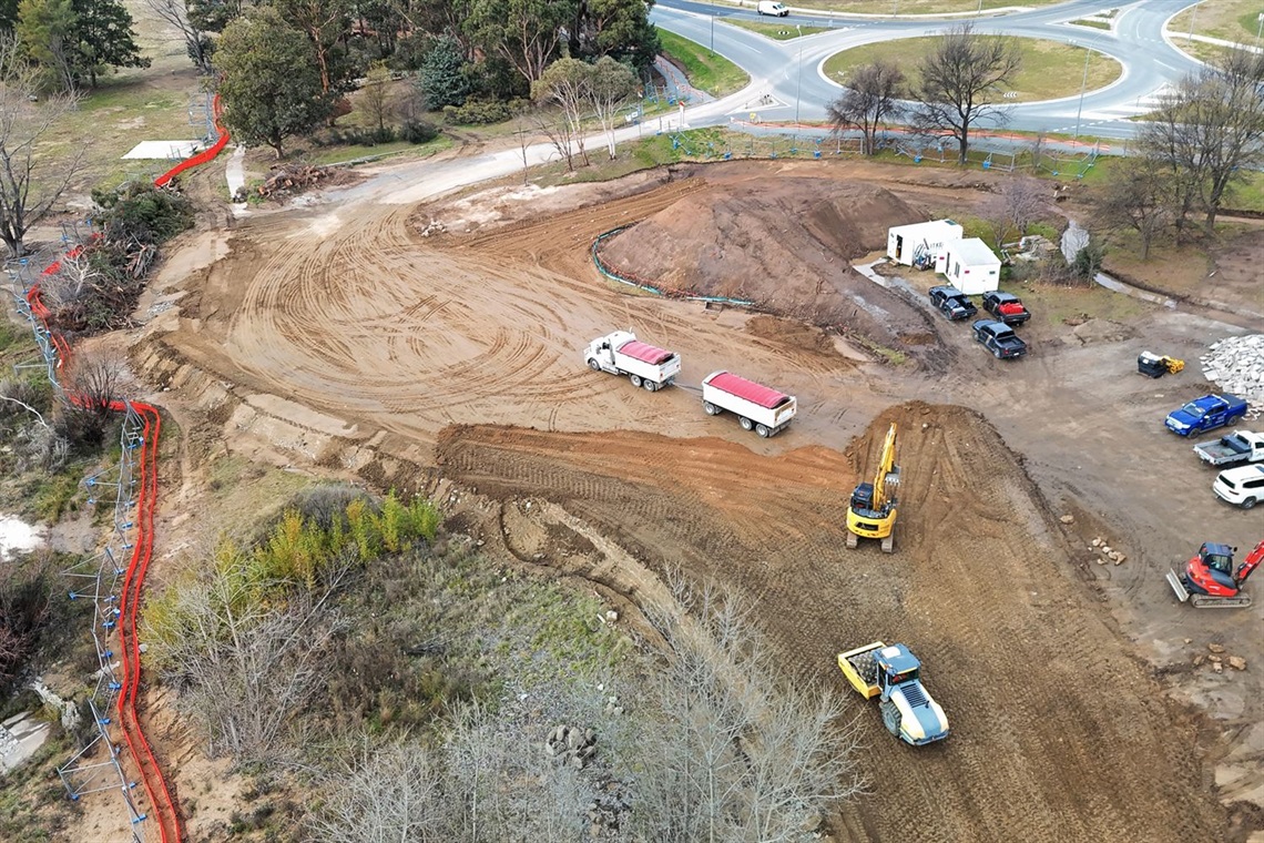 Earthworks seen from above, with construction vehicles, a large truck and a number of utes, with trees, shrubs and orange barrier fencing surrounding.