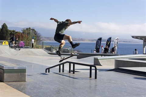 A skateboarder mid-kickflip in the air above a rail in the street terrain section of the new Jindabyne Skate Park, with the Lake and attendees in the background.