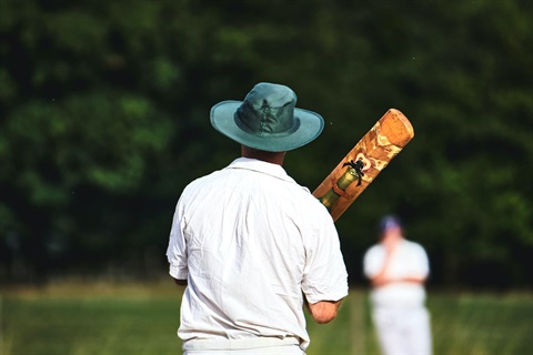A man in cricketing whites and green wide-brimmed hat from behind, holding a cricket bat over his shoulder outdoors.