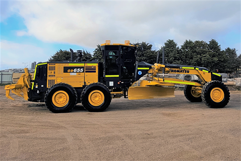 A large bright yellow and black Komatsu-brand road grader parked on a gravel lot, seen from the side.