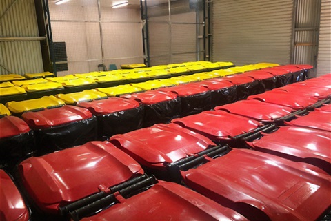 Rows of red and yellow-lidded domestic wheelie bins inside a concrete and steel structure.