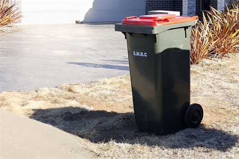Red-lid kerbisde rubbish bin sitting on kerb outside suburban home