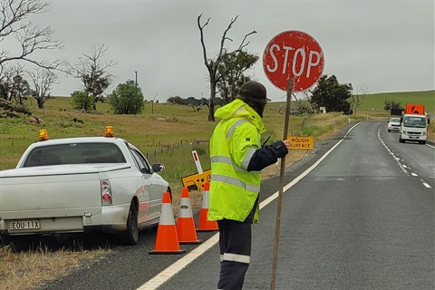 A traffic controller in a high visibility raincoat holds a stop sign standing on a two-lane road on an overcast day, a work truck is in the background and a ute to his left