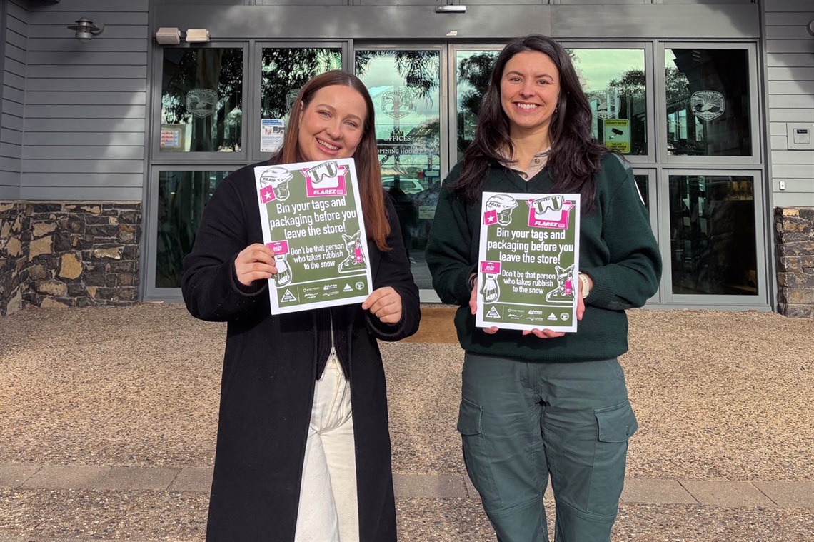 Two women smiling, posing with colourful anti-littering campaign posters at the entrance to a stone-and-brick building exterior