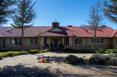 A single-storey blonde-brick building surrounded by foliage, garden beds and paths with a red metal roof