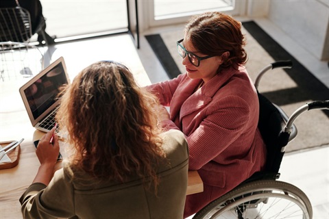 Two women in front of a laptop computer, one in a wheelchair