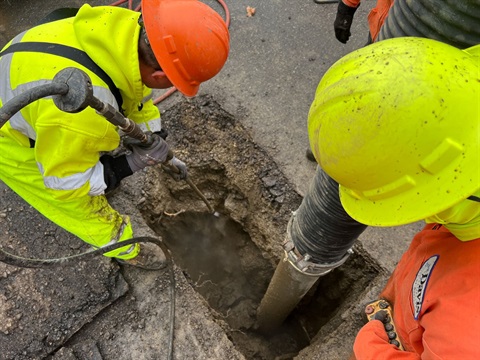 Workers in high vis jumpers and hardhats repairing a water main in a wet, freshly dug trench.